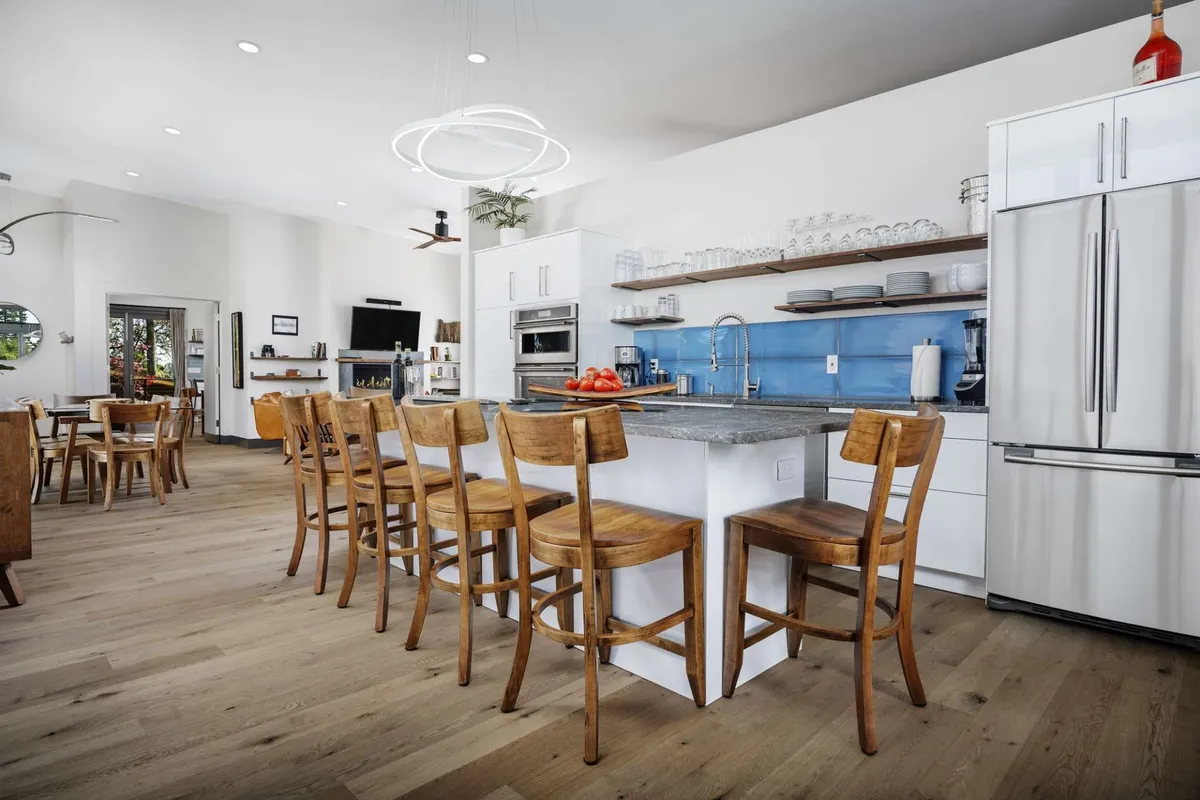 A bright and modern gourmet kitchen with a central island, bar stools, a blue tiled backsplash, and wooden dining chairs. The setup invites guests to share casual meals, prepare food together, or enjoy conversations over coffee under natural daylight.