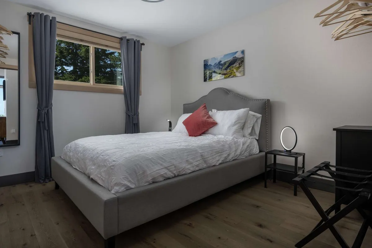 This photo shows a serene bedroom with a large window allowing ample natural light, a bed with white bedding and a pop of color from a red accent pillow, minimalist decor, and calming artwork. The space exudes tranquility and comfort, ideal for restful sleep.