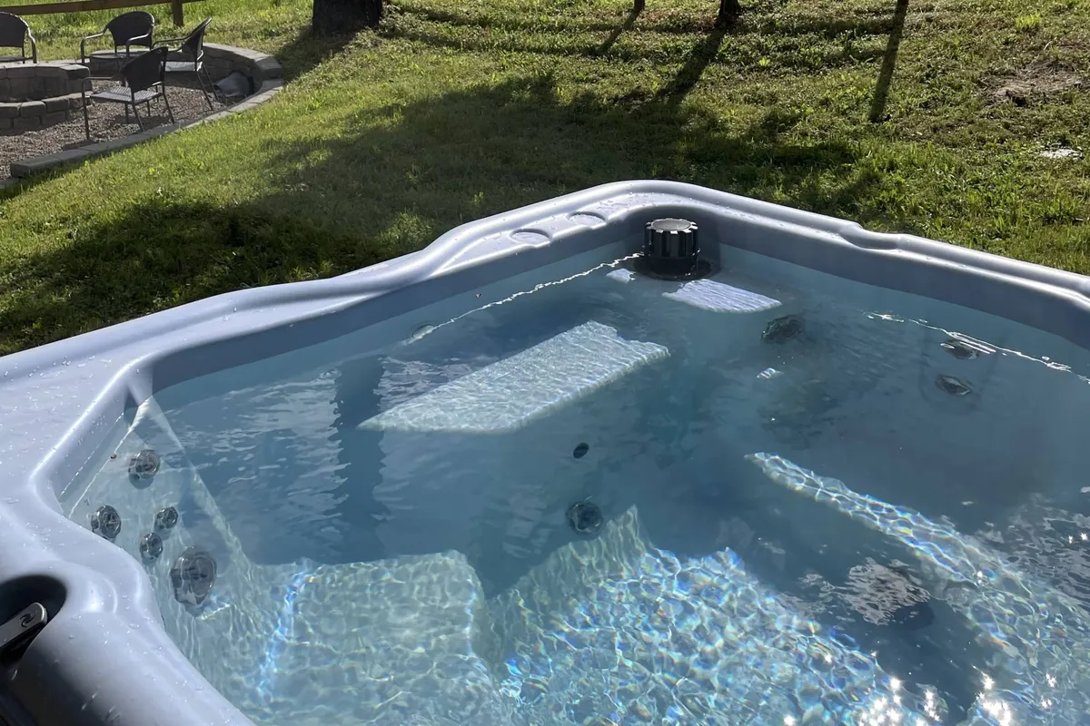 A tranquil exterior scene with a brand-new hot tub set atop lush grass, adjacent to a cozy seating area. The hot tub offers stunning sunset reflections on the water, emphasizing both relaxation and breathtaking natural surroundings.