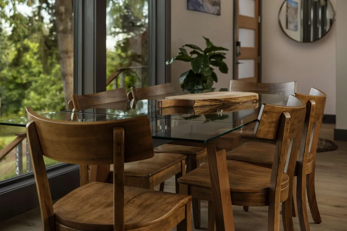 A glass dining table surrounded by wooden chairs, set beside large windows and complemented by an indoor plant and decorative mirror. The space is arranged for group meals, game nights, or collaborative work sessions, with plenty of natural light and a welcoming, open feel.