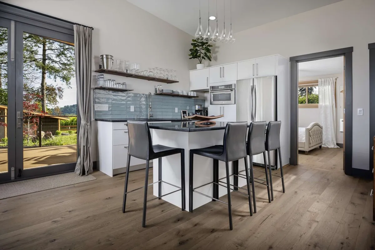 This image displays a sleek, well-equipped kitchenette with a kitchen island, contemporary appliances, glassware on open shelves, and expansive windows that create a welcoming, airy atmosphere. The space is ideal for preparing meals, entertaining, or casual breakfasts.