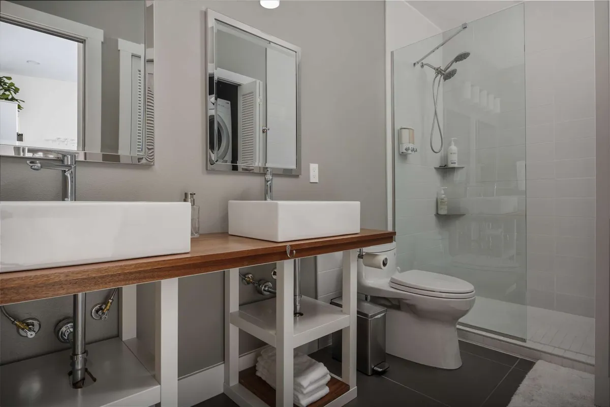 A contemporary bathroom with a double sink vanity, glass shower enclosure, and mirrors. The space is bright, clean, and spacious, offering both luxury and privacy for guests in each wing.