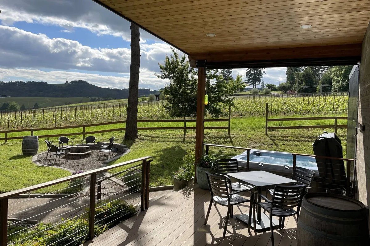 A welcoming patio area with a bubbling jacuzzi, a cozy fire pit surrounded by seating, and sweeping vineyard views. The space is framed by large shade trees, offering an elegant blend of privacy and connection to nature.