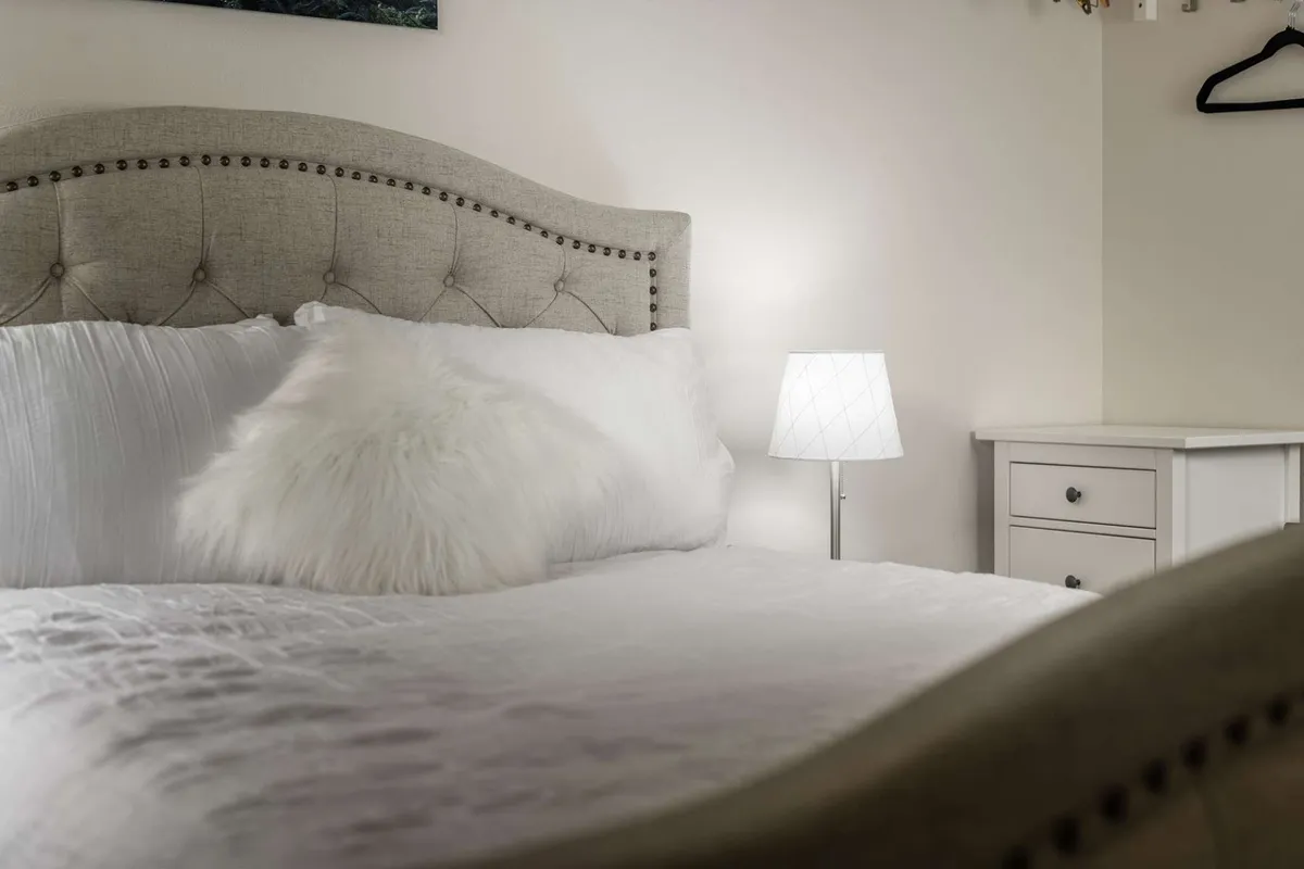 A tranquil bedroom featuring a tufted headboard, fluffy white pillow, and a nightstand with a lamp. The soft lighting and high-quality bedding create a serene and inviting atmosphere, perfect for restful nights.