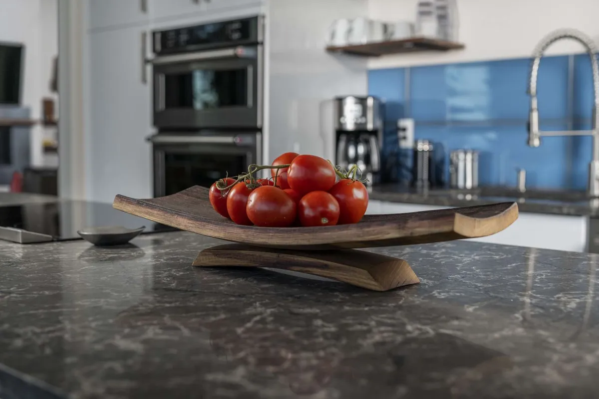 A view of the kitchen featuring sleek appliances, a marble countertop, and a wooden bowl of red tomatoes, all set up for culinary activity and communal gathering. The space feels warm and ready for shared meals or family feasts.
