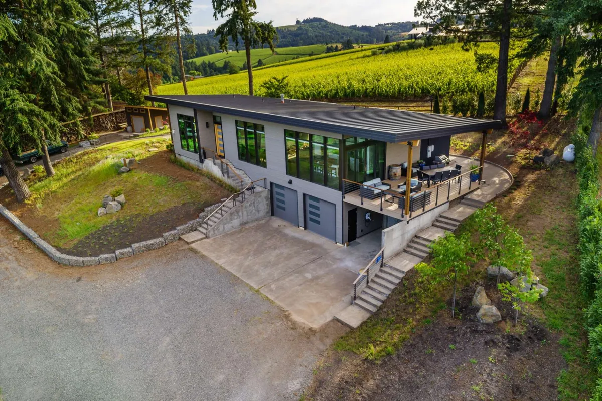 The image showcases the villa’s striking modern architecture set amid lush, landscaped gardens. Expansive floor-to-ceiling windows frame the sunset and fill the interior with golden light. A spacious outdoor seating and dining area invites guests to gather, relax, and savor panoramic views of Oregon wine country.