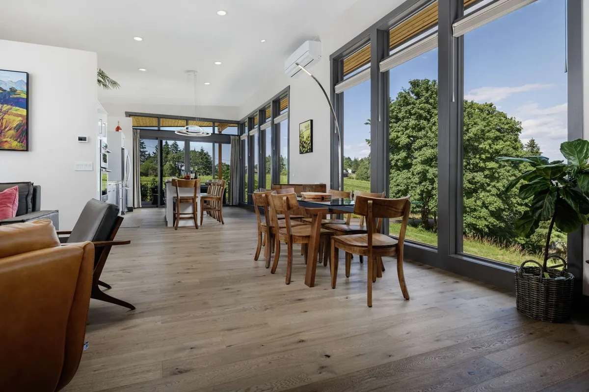 A spacious living and dining area bathed in natural light from expansive floor-to-ceiling windows, offering sweeping views of lush greenery and the surrounding landscape. The scene includes a large table with chairs, a plush seating area, and sunlight streaming in, creating an inviting spot for both lively gatherings and quiet moments.