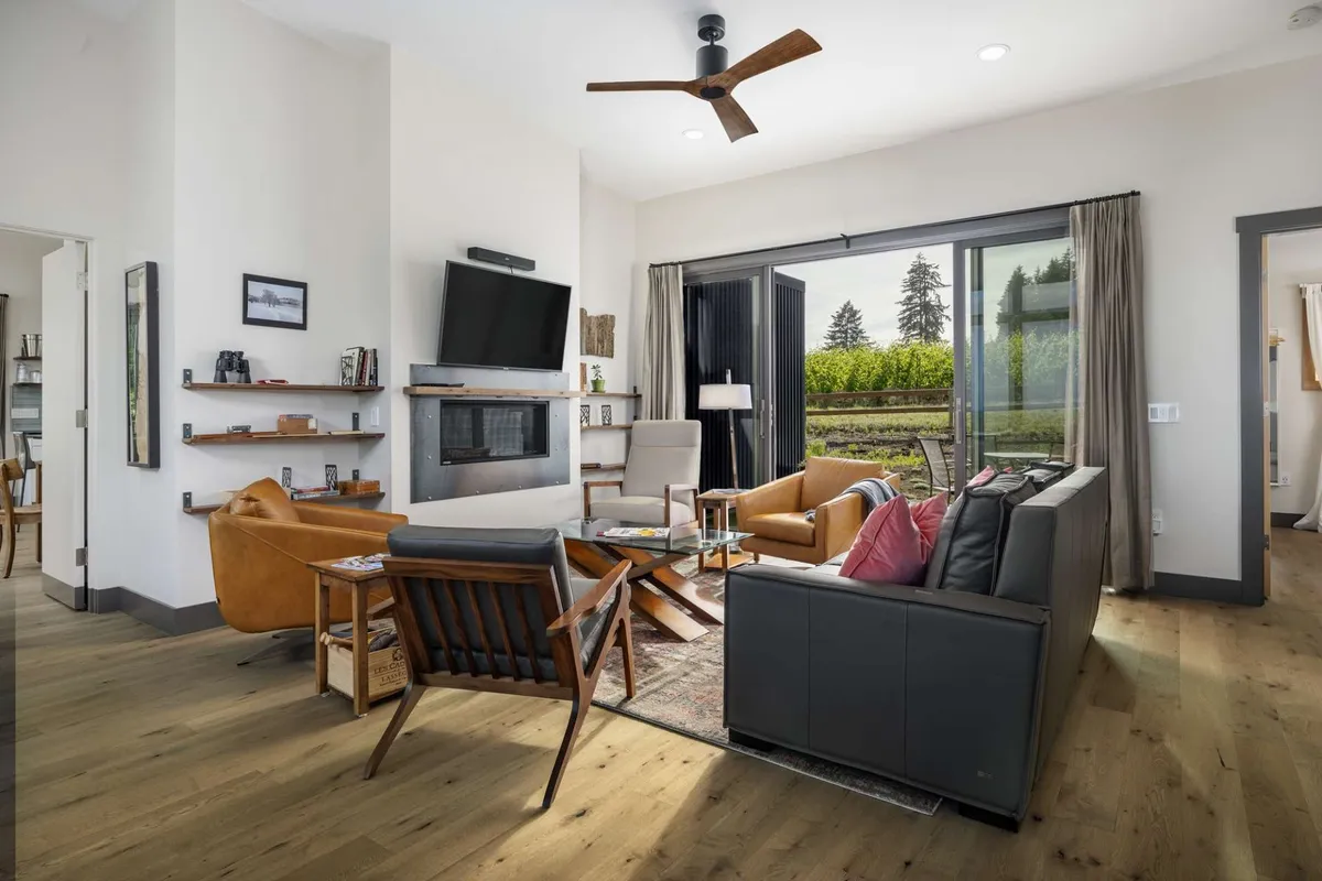 A cozy living room with large windows showcasing vineyard views, a modern fireplace, comfortable seating, stylish décor, books, and art. The space feels both sophisticated and inviting, ideal for relaxing with loved ones in front of the fire as the sun sets outside.
