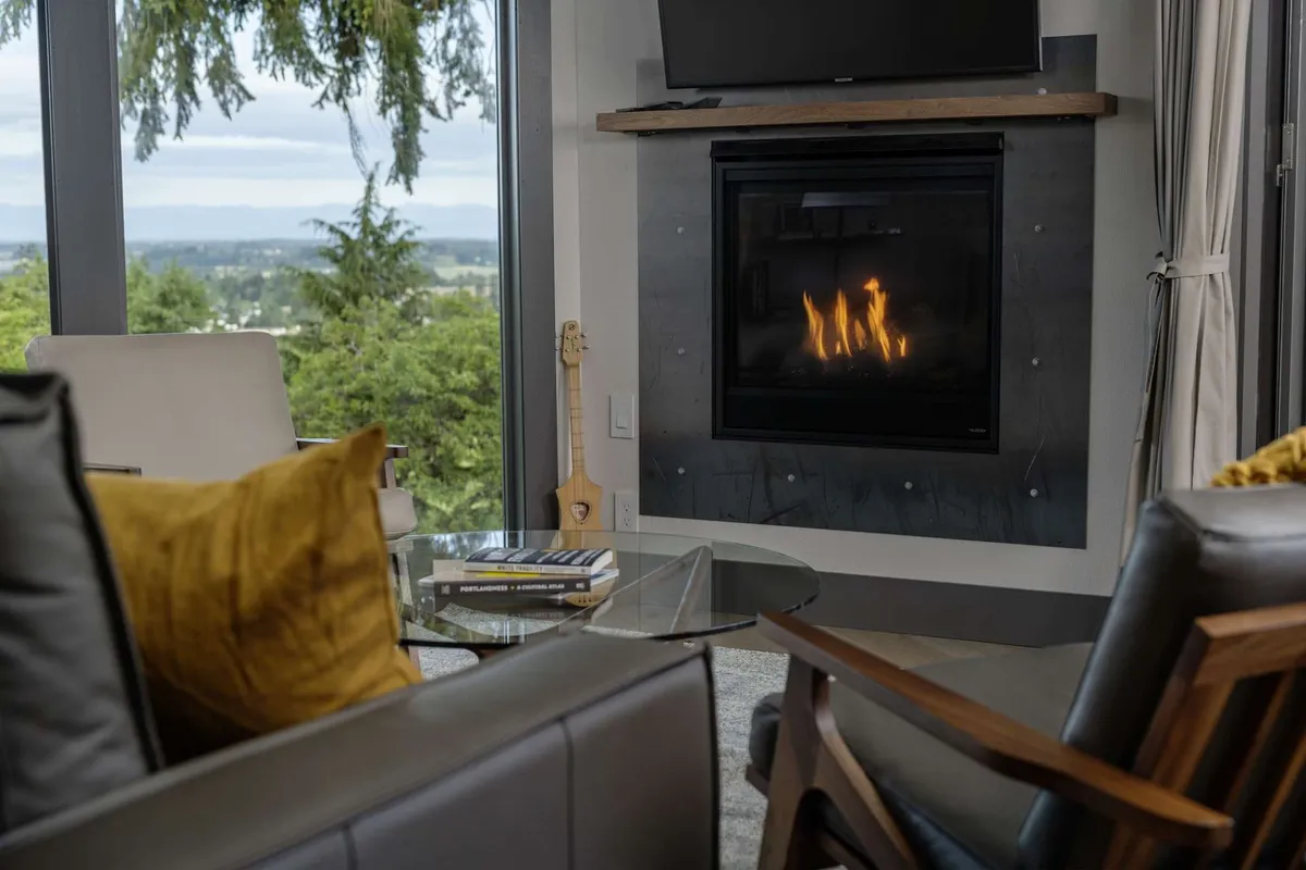 A spacious living area with a modern fireplace, large windows framing valley views, and a cozy seating arrangement around a coffee table. The room is filled with natural light, offering an inviting space for group discussions, casual gatherings, or quiet moments of inspiration. The atmosphere is both warm and professional, suited for both work and relaxation.
